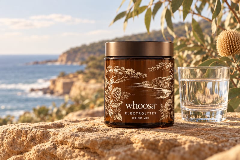 Concept render of the WHOOSA Electrolytes drink mix: an amber wide-mouth canister with a bronze-coloured lid, labelled ELECTROLYTES DRINK MIX, next to a glass of water on sandstone