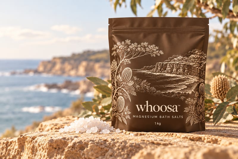 Concept render of the WHOOSA Magnesium Bath Salts in a bronze-brown stand-up resealable pouch with ivory coastal linework, marked 1 kg, beside a small pile of salt crystals on sandstone